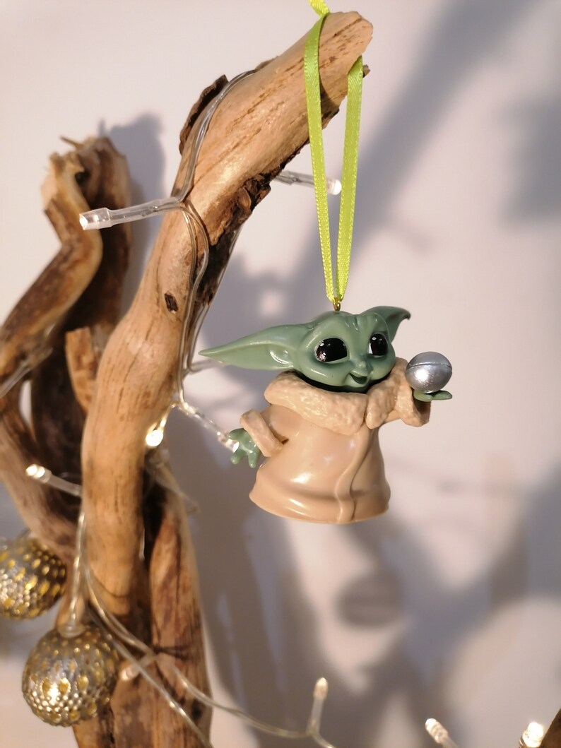 May include: A green and brown Christmas ornament shaped like a character from the Star Wars franchise. The ornament is holding a silver ball in its hand.