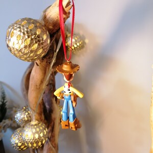 May include: A red string hangs from a wooden branch with a small plastic toy of Woody from Toy Story hanging from it. The toy is wearing his signature cowboy hat and vest. The branch is decorated with silver and white string lights.