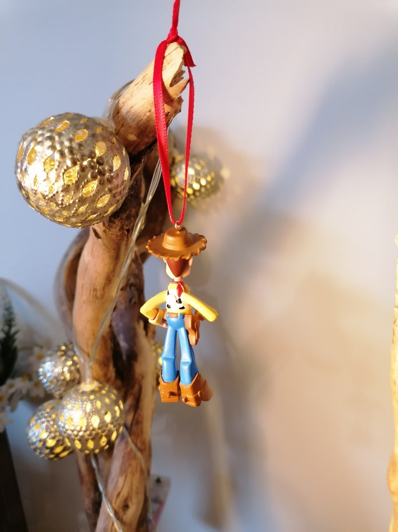 May include: A red string hangs from a wooden branch with a small plastic toy of Woody from Toy Story hanging from it. The toy is wearing his signature cowboy hat and vest. The branch is decorated with silver and white string lights.