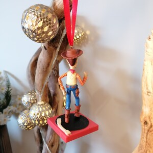 Puede incluir: Un adorno navide&ntilde;o rojo y negro con una figura de pl&aacute;stico de Woody de Toy Story de pie sobre un tocadiscos rojo y negro.