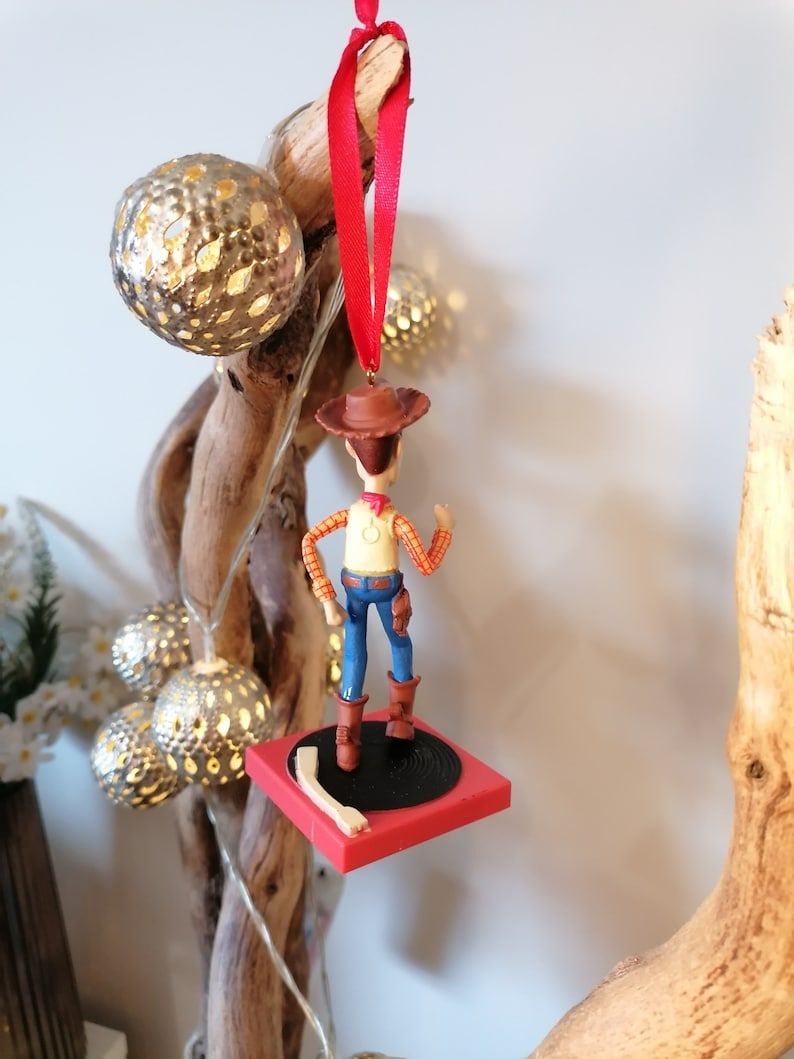 Puede incluir: Un adorno navide&ntilde;o rojo y negro con una figura de pl&aacute;stico de Woody de Toy Story de pie sobre un tocadiscos rojo y negro.