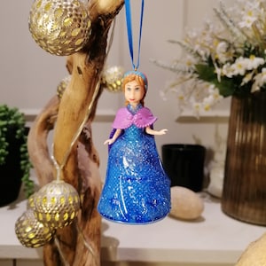 Puede incluir: Un adorno de pl&aacute;stico azul y morado de una princesa con un vestido azul con purpurina. El adorno est&aacute; colgado de una cinta azul.