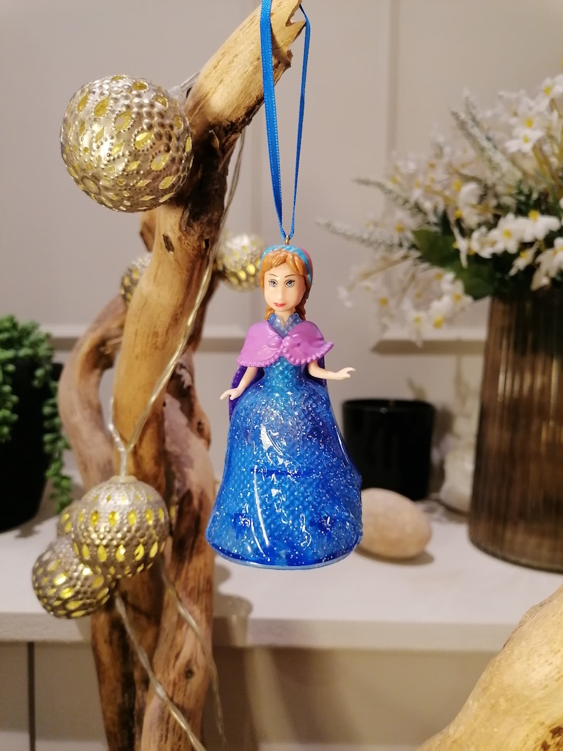 Puede incluir: Un adorno de pl&aacute;stico azul y morado de una princesa con un vestido azul con purpurina. El adorno est&aacute; colgado de una cinta azul.