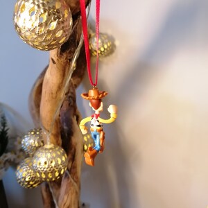 May include: A red string ornament featuring a plastic figurine of Woody from the Toy Story franchise. Woody is wearing his signature cowboy hat, vest, and jeans. He is holding his lasso and has a friendly expression on his face.