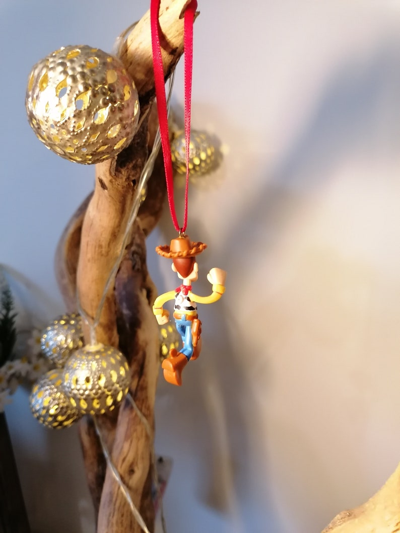 May include: A red string ornament featuring a plastic figurine of Woody from the Toy Story franchise. Woody is wearing his signature cowboy hat, vest, and jeans. He is holding his lasso and has a friendly expression on his face.