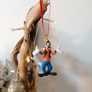 Op de afbeelding: Een rode touwversiering met een cartoonfiguur van Goofy met een gele hoed, een oranje shirt, een blauwe broek en een zwart vest. Goofy glimlacht en steekt zijn tong uit.
