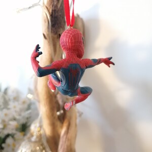 May include: Red and blue Spider-Man Christmas ornament hanging from a red ribbon. The ornament is a plastic figurine of Spider-Man in a web-slinging pose.