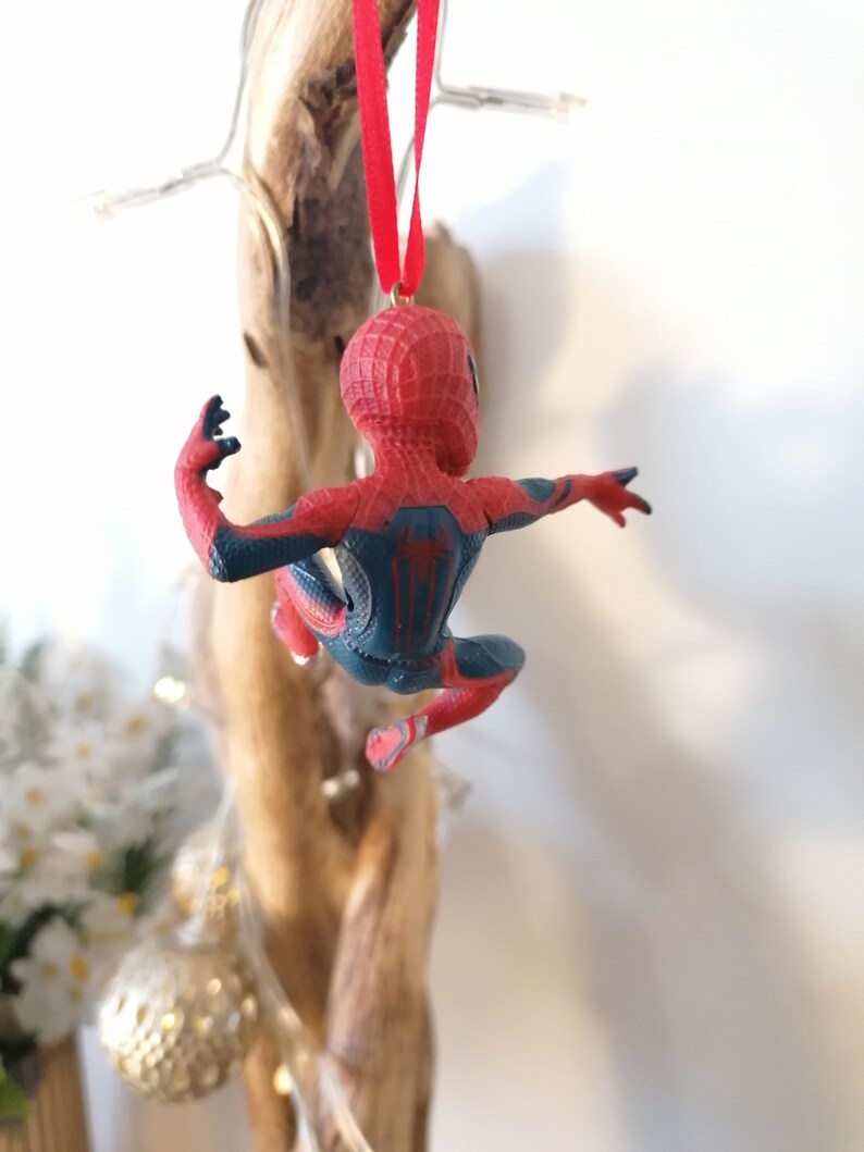 May include: Red and blue Spider-Man Christmas ornament hanging from a red ribbon. The ornament is a plastic figurine of Spider-Man in a web-slinging pose.