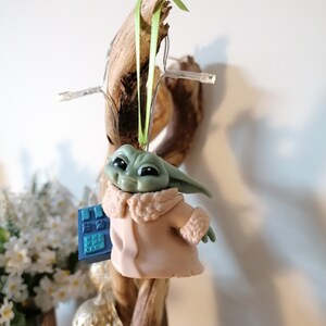 May include: A green and brown figurine of a character from the Star Wars franchise, wearing a pink coat with a white fur trim. The figurine is holding a blue device and is hanging from a green ribbon.