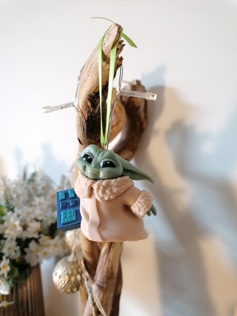 May include: A green and brown figurine of a character from the Star Wars franchise, wearing a pink coat with a white fur trim. The figurine is holding a blue device and is hanging from a green ribbon.