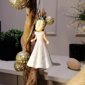 Peut inclure: Une d&eacute;coration en plastique blanche et bleue repr&eacute;sentant une princesse portant une robe blanche avec une ceinture bleue. La d&eacute;coration est suspendue &agrave; une branche.