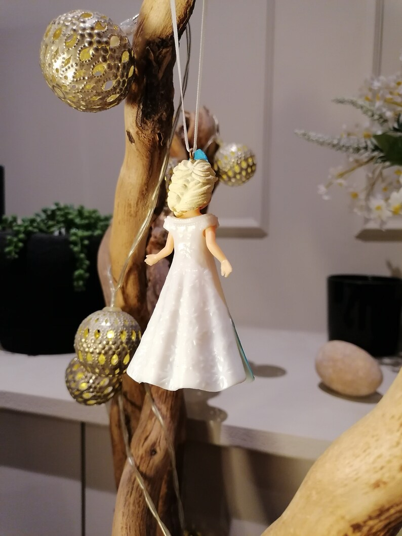 Peut inclure: Une d&eacute;coration en plastique blanche et bleue repr&eacute;sentant une princesse portant une robe blanche avec une ceinture bleue. La d&eacute;coration est suspendue &agrave; une branche.