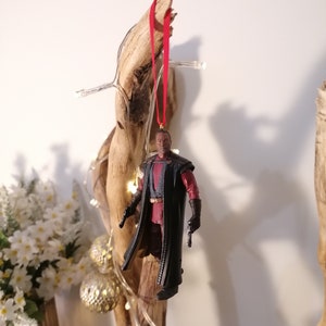 K&ouml;nnte beinhalten: Eine schwarze und rote Actionfigur eines Charakters, der einen langen schwarzen Mantel tr&auml;gt und zwei Waffen h&auml;lt. Die Figur h&auml;ngt an einem roten Band an einem St&uuml;ck Treibholz.