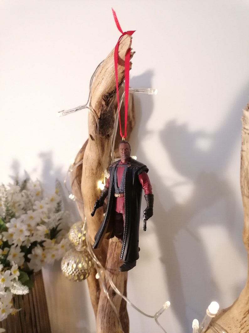 K&ouml;nnte beinhalten: Eine schwarze und rote Actionfigur eines Charakters, der einen langen schwarzen Mantel tr&auml;gt und zwei Waffen h&auml;lt. Die Figur h&auml;ngt an einem roten Band an einem St&uuml;ck Treibholz.