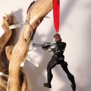 May include: A black and silver action figure of a superhero holding a gun, hanging from a red ribbon. The figure is wearing a black suit with silver accents and a black helmet. The figure is posed in a dynamic action pose.