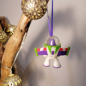 Puede incluir: Un adorno navide&ntilde;o de Buzz Lightyear de color morado y verde colgado de una rama con una cinta morada. El adorno es una peque&ntilde;a figura de pl&aacute;stico de Buzz Lightyear de las pel&iacute;culas de Toy Story.
