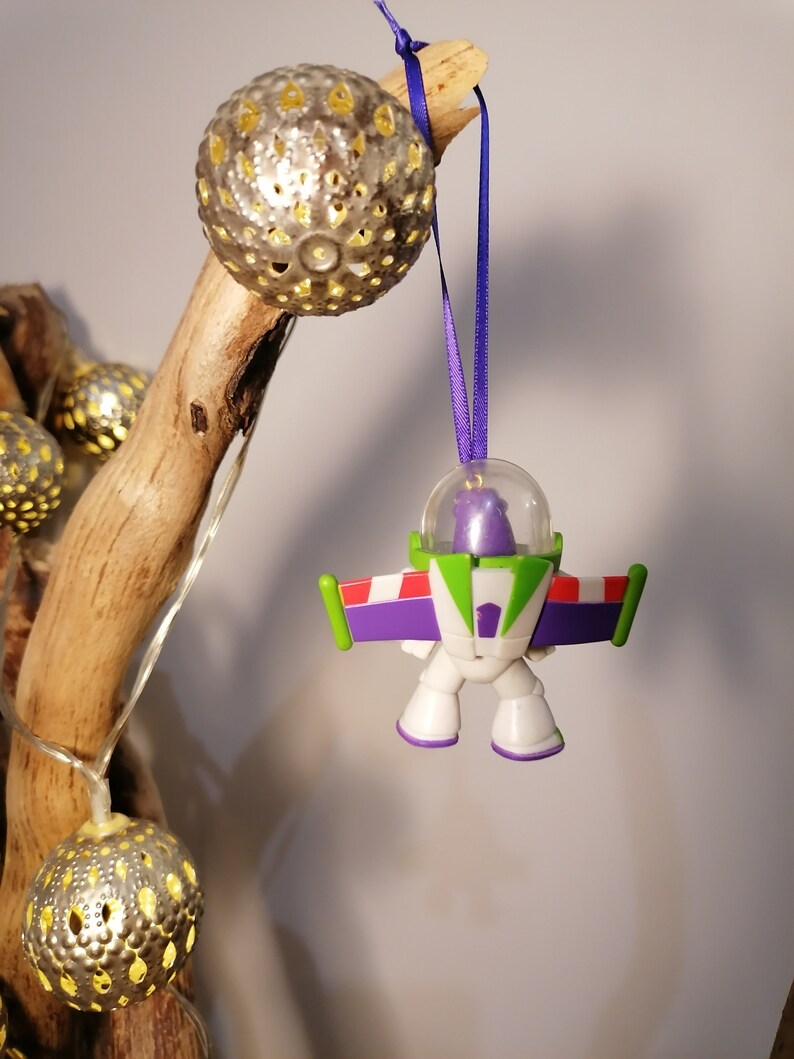 Puede incluir: Un adorno navide&ntilde;o de Buzz Lightyear de color morado y verde colgado de una rama con una cinta morada. El adorno es una peque&ntilde;a figura de pl&aacute;stico de Buzz Lightyear de las pel&iacute;culas de Toy Story.