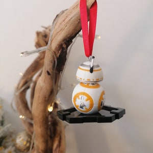 May include: A white and orange Star Wars BB-8 droid ornament with a black stand, hanging from a red ribbon on a wooden branch.