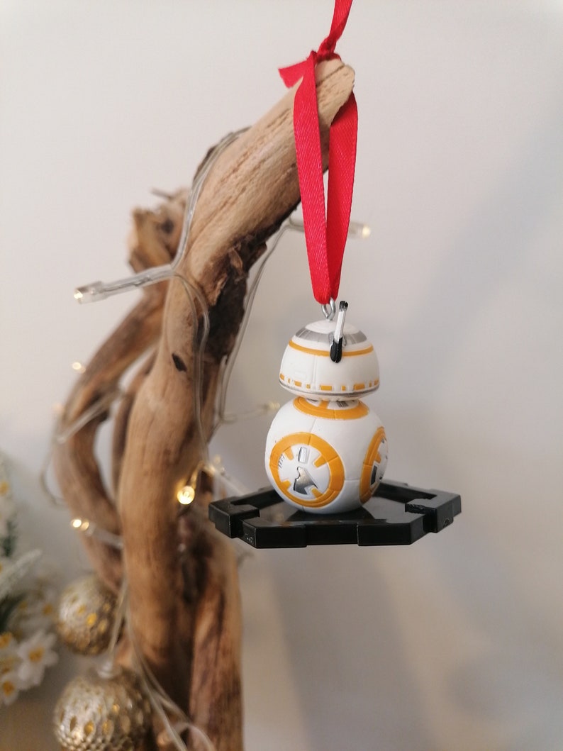 May include: A white and orange Star Wars BB-8 droid ornament with a black stand, hanging from a red ribbon on a wooden branch.
