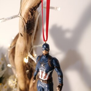 May include: A blue and red Captain America action figure ornament with a red ribbon hanging from the top. The figure is wearing a blue and red suit with a white star on the chest and a red, white, and blue shield on his arm.
