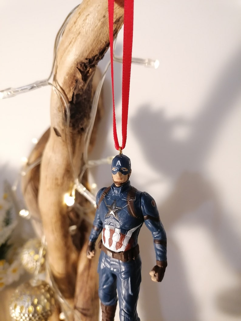 May include: A blue and red Captain America action figure ornament with a red ribbon hanging from the top. The figure is wearing a blue and red suit with a white star on the chest and a red, white, and blue shield on his arm.