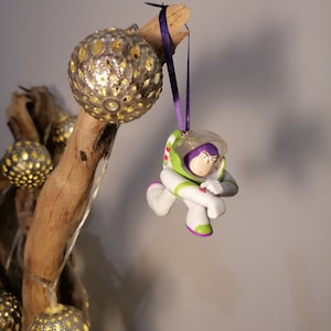 Puede incluir: Un adorno de figura de acción de ranger espacial verde y blanco colgando de una cinta morada en una rama de madera a la deriva con adornos plateados y dorados.