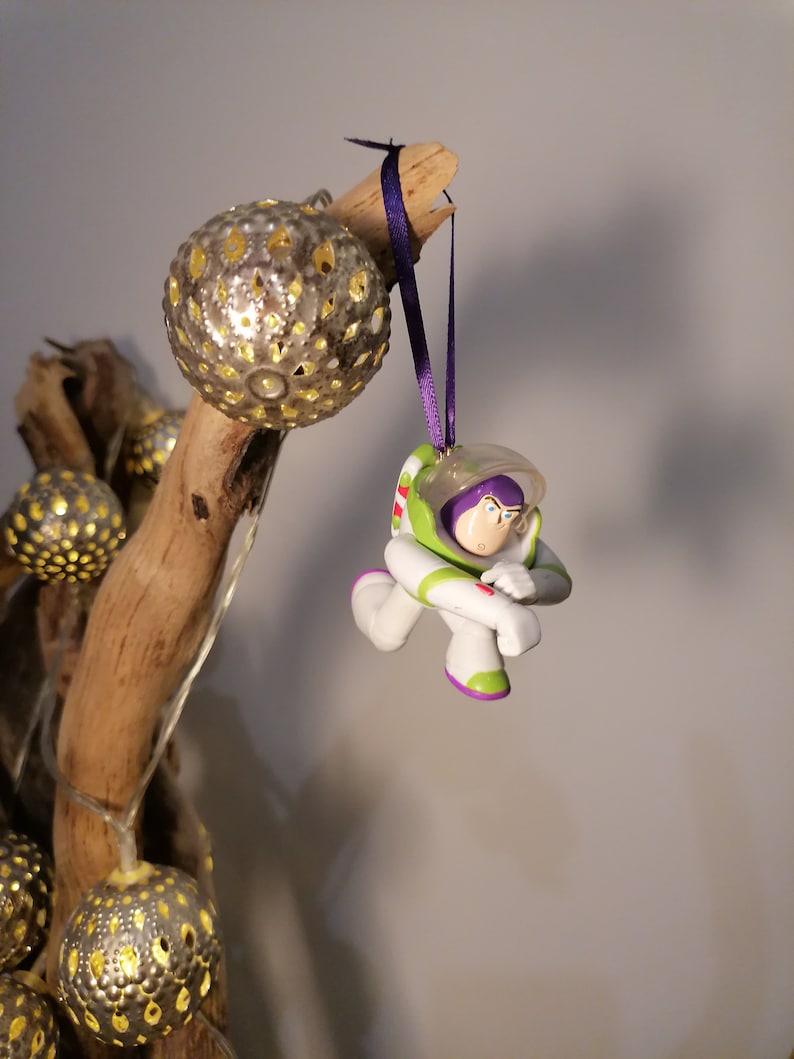 Puede incluir: Un adorno de figura de acci&oacute;n de ranger espacial verde y blanco colgando de una cinta morada en una rama de madera a la deriva con adornos plateados y dorados.