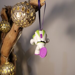 Puede incluir: Un adorno navide&ntilde;o de Buzz Lightyear verde, blanco y morado colgado de una rama con una cinta morada. El adorno es una versi&oacute;n peque&ntilde;a y estilizada del personaje de las pel&iacute;culas Toy Story.