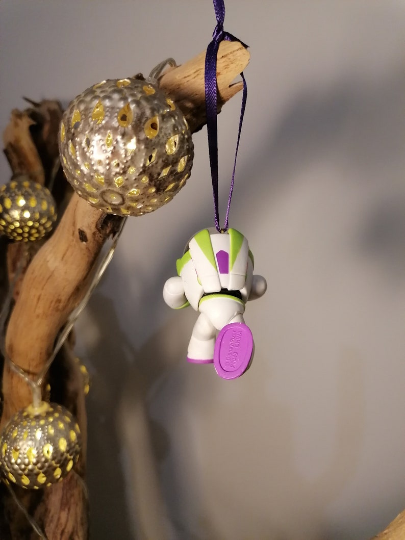 Puede incluir: Un adorno navide&ntilde;o de Buzz Lightyear verde, blanco y morado colgado de una rama con una cinta morada. El adorno es una versi&oacute;n peque&ntilde;a y estilizada del personaje de las pel&iacute;culas Toy Story.