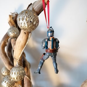 May include: A blue and brown Star Wars Boba Fett Christmas ornament hanging from a red ribbon on a branch with silver ornaments.