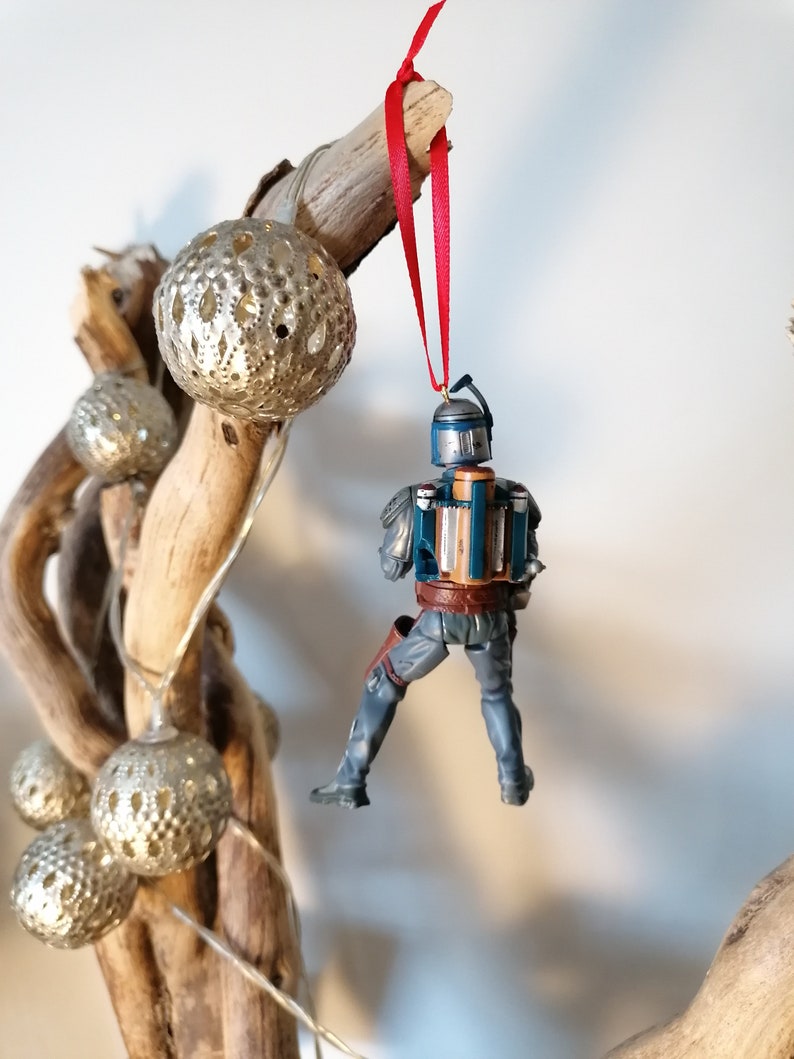 May include: A blue and brown Star Wars Boba Fett Christmas ornament hanging from a red ribbon on a branch with silver ornaments.