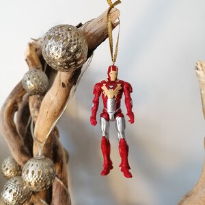Pode incluir: Ornamento de figura de a&ccedil;&atilde;o do Homem de Ferro vermelho e prateado pendurado em um galho de madeira flutuante com um cord&atilde;o dourado e bolas de glitter douradas.