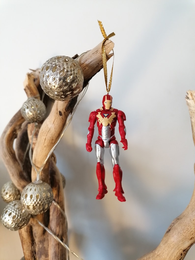 Pode incluir: Ornamento de figura de a&ccedil;&atilde;o do Homem de Ferro vermelho e prateado pendurado em um galho de madeira flutuante com um cord&atilde;o dourado e bolas de glitter douradas.