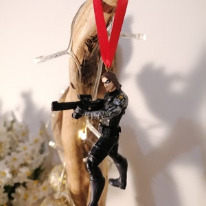 May include: A black and gray action figure of a superhero holding a gun, wearing a black and gray suit and a black mask. The figure is hanging from a red ribbon attached to a piece of driftwood.