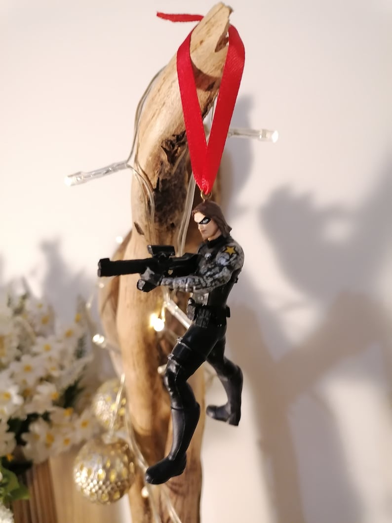 May include: A black and gray action figure of a superhero holding a gun, wearing a black and gray suit and a black mask. The figure is hanging from a red ribbon attached to a piece of driftwood.
