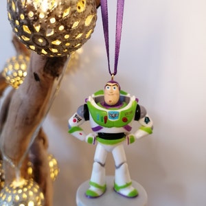 May include: A green and white plastic ornament of Buzz Lightyear from the Toy Story franchise. The ornament is hanging from a purple ribbon and has a gold hook.