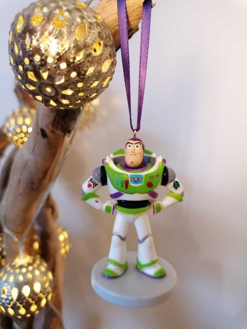 May include: A green and white plastic ornament of Buzz Lightyear from the Toy Story franchise. The ornament is hanging from a purple ribbon and has a gold hook.