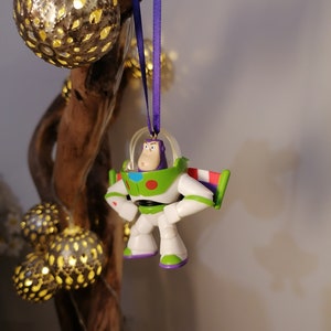 Puede incluir: Un adorno navide&ntilde;o de Buzz Lightyear blanco y verde con una cinta morada colgando de una rama. El adorno es una peque&ntilde;a figura de pl&aacute;stico del personaje de las pel&iacute;culas Toy Story.