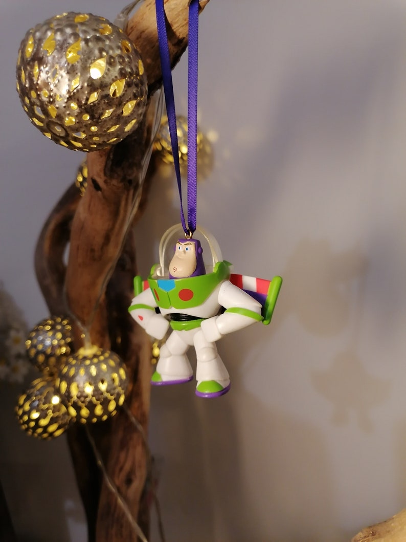 Puede incluir: Un adorno navide&ntilde;o de Buzz Lightyear blanco y verde con una cinta morada colgando de una rama. El adorno es una peque&ntilde;a figura de pl&aacute;stico del personaje de las pel&iacute;culas Toy Story.