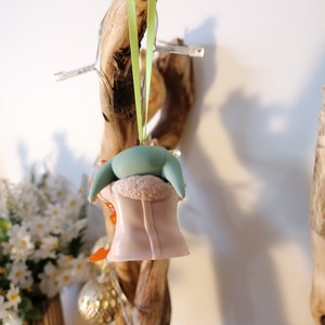 May include: A green and pink Christmas bauble shaped like a character from the Star Wars franchise. The bauble is hanging from a green ribbon on a wooden branch.