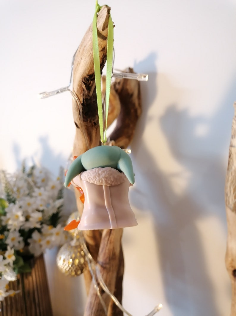 May include: A green and pink Christmas bauble shaped like a character from the Star Wars franchise. The bauble is hanging from a green ribbon on a wooden branch.