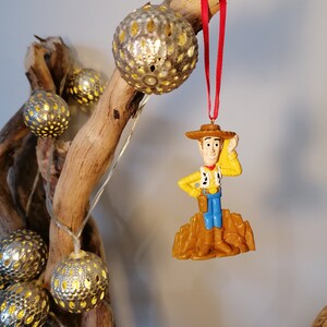 May include: A plastic ornament of Woody from Toy Story, wearing his signature cowboy hat and vest, standing on a brown base. The ornament is hanging from a red ribbon on a branch with string lights.