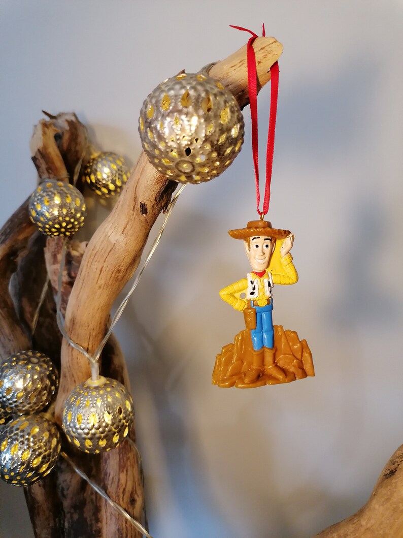 May include: A plastic ornament of Woody from Toy Story, wearing his signature cowboy hat and vest, standing on a brown base. The ornament is hanging from a red ribbon on a branch with string lights.