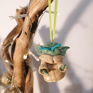 May include: A green and brown Yoda ornament with a blue lightsaber hanging from a green ribbon on a branch with white lights.