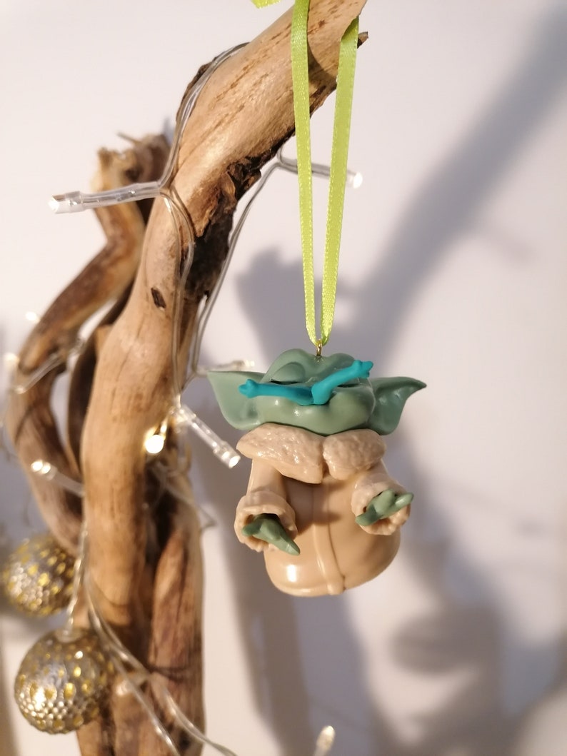 May include: A green and brown Yoda ornament with a blue lightsaber hanging from a green ribbon on a branch with white lights.