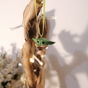 May include: A green and brown figurine of a character from the Star Wars franchise, hanging from a green ribbon on a piece of driftwood. The figurine is holding a small brown object.