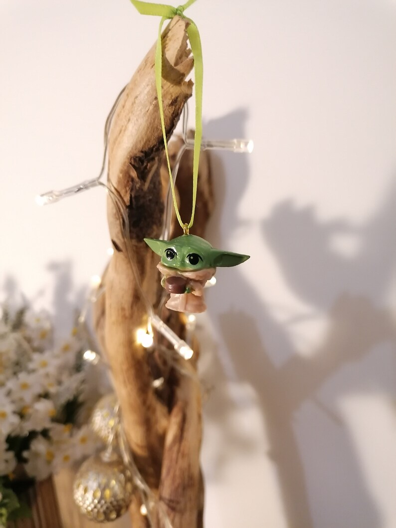 May include: A green and brown figurine of a character from the Star Wars franchise, hanging from a green ribbon on a piece of driftwood. The figurine is holding a small brown object.