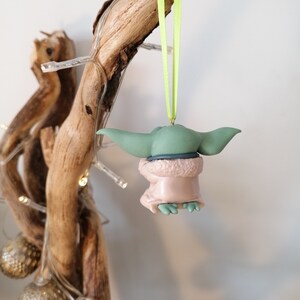 May include: A green and brown Christmas ornament shaped like a character from the Star Wars franchise. The ornament is hanging from a green ribbon.