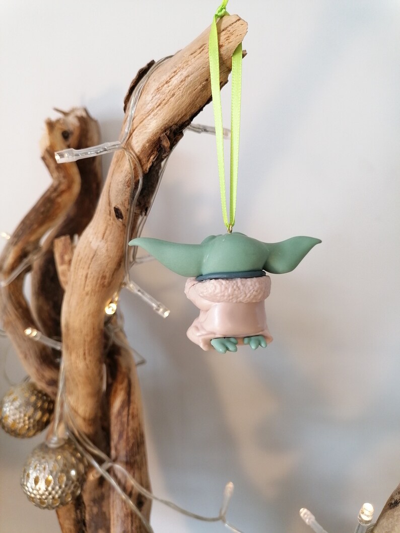 May include: A green and brown Christmas ornament shaped like a character from the Star Wars franchise. The ornament is hanging from a green ribbon.