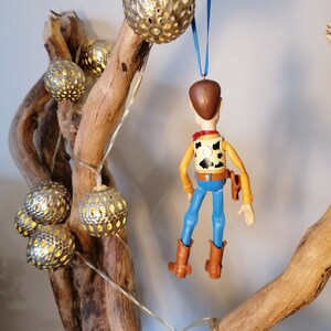Puede incluir: Una rama de madera decorada con una cadena de luces y un adorno colgante de una figura de vaquero de juguete. Las luces son redondas y tienen un acabado met&aacute;lico con dise&ntilde;os recortados. La figura de vaquero lleva un chaleco marr&oacute;n, pantalones vaqueros azules y un sombrero marr&oacute;n.